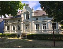 Eine weiße Villa mit reich verzierten Fassaden, hohen Fenstern und einem kleinen Eingangsvorbau mit Treppe. Der Vordergarten ist von Hecken umgeben, und große Bäume spenden teilweise Schatten.