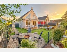 Ein modernes Einfamilienhaus im skandinavischen Stil mit einem spitzen Giebeldach und großen Glasfenstern, umgeben von einem gepflegten Garten mit Gartenmöbeln, einer Feuerstelle und einer Holzsauna im Hintergrund. Der Sonnenuntergang taucht die Szene in warmes Licht.