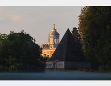 Eine Landschaft mit einer steinernen Pyramide im Vordergrund und einem historischen Gebäude mit Kuppel und Verzierungen im Hintergrund, umgeben von Bäumen und leichtem Nebel in der Dämmerung.