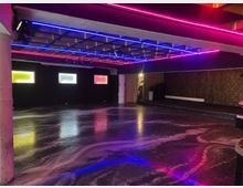 Großer, leerer Innenraum wie ein Club- oder Veranstaltungsraum mit glänzendem dunklem Boden und einer Decke mit pinken und blauen Neon-Lichtleisten. An der Rückwand befinden sich drei kleine rechteckige Fenster mit farbigem Licht, daneben eine Tür und an der rechten Wand eine lange niedrige Sitzbank vor gemusterter Tapete.