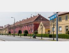 Straßenszene mit einer Reihe von Wohnhäusern in gelbem und rotem Putz sowie roten Ziegeldächern; im Vordergrund stehen kleine Straßenbäume und eine grüne Motorradmaschine auf dem Gehweg. Ein Straßenschild mit der Aufschrift „Jüterboger Tor“ ist ebenfalls sichtbar.