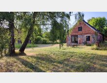 Ein Backsteingebäude mit einem Dach aus Schiefer und einer Fassade aus teilweise freiliegenden Steinen, umgeben von einer Wiese mit Bäumen. Im Vordergrund stehen Gartenmöbel, darunter ein Holztisch und Stühle, auf einem gepflegten Grünbereich.