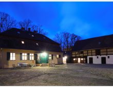 Hof mit historischen Gebäuden bei Dämmerung, bestehend aus einem gelb gestrichenen Haus mit beleuchteten Fenstern und grüner Eingangstür sowie einem Fachwerkgebäude. Der gepflasterte Hof ist umgeben von Bäumen, die sich im Hintergrund abzeichnen.
