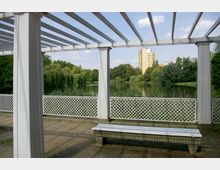 Weiße Pergola mit Gitterzaun und einer Bank im Vordergrund, Blick auf einen Park mit einem Teich, Weidenbäumen und einem gelben Hochhaus im Hintergrund.