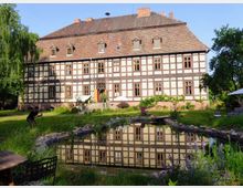 Historisches Fachwerkgebäude mit rotem Ziegeldach, umgeben von einem Garten mit einem Teich im Vordergrund. Ein Pfad führt von einer Terrasse mit Sonnenschirmen zum Wasser, während Bäume und Pflanzen die Landschaft ergänzen.