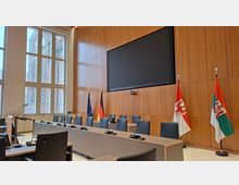 Moderner Konferenz- oder Sitzungssaal mit langen Tischen, grauen Stühlen und integrierten Mikrofonen, vor einer holzvertäfelten Wand mit großem Bildschirm. Seitlich stehen hohe Fenster, und neben dem Podium sind mehrere Flaggen (EU, Deutschland sowie Landesflaggen) aufgestellt.