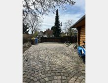 Hof mit gepflastertem Boden aus unregelmäßigen Steinen, umgeben von Bäumen und einem Holzzaun. Rechts im Bild befindet sich ein blauer Motorroller, ein Fahrrad und ein Holzgebäude, während links Mülltonnen an der Seite stehen.