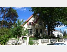 Gelbes Einfamilienhaus mit rotem Satteldach und Fensterläden, umgeben von Bäumen und einem Garten. Vor dem Haus steht ein weißer Zaun und ein Auto ist im Vordergrund geparkt.