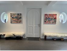 Eingangsbereich mit einer weißen Tür in der Mitte, flankiert von zwei runden Fenstern. An den Wänden hängen bunte Gemälde, unter den Fenstern stehen Holzbänke mit weißen Schaffellen, darunter Schuhablagen.