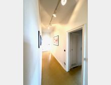 Langer, schmaler Innenflur mit weißen Wänden, glattem Boden und Deckenbeleuchtung über einer Schiene. An den Wänden hängen gerahmte Bilder, rechts öffnet sich eine Tür zu einem angrenzenden Raum mit einer weiteren weißen Tür.