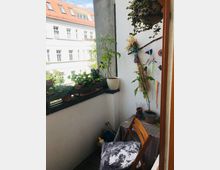 Ein kleiner Balkon mit weißen Wänden und einem Tisch mit gemusterter Tischdecke, darauf Pflanzen in Töpfen. An der Wand hängen dekorative Elemente, und auf der Brüstung stehen weitere Pflanzen. Im Hintergrund ist die Fassade eines Wohnhauses mit roten Dachziegeln zu sehen.