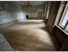 Ein leerer Raum mit Holzdielenboden, abgeblätterter Wandfarbe und hohen Fenstern mit Holzrahmen auf der rechten Seite. Eine einfache Holztür sowie ein Kamin befinden sich an der gegenüberliegenden Wand, und eine dekorative Hängelampe ist an der Decke angebracht.