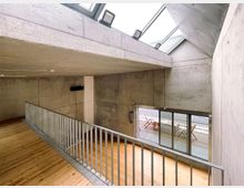 Interior of a modern concrete building with a wooden floor walkway and metal railing overlooking a lower level. Large skylights and a wall of sliding glass doors open to a small outdoor deck with seating.