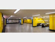 U-Bahn-Station mit gelb verkleideten Wänden und Decken, beleuchtet durch zahlreiche Leuchtstoffröhren. Sichtbar sind Rolltreppen auf der rechten Seite sowie Infotafeln und Werbeflächen an den Wänden.