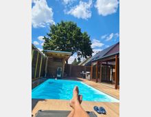 Privater Garten mit einem rechteckigen Swimmingpool, umgeben von Holzdielen. Im Hintergrund befinden sich ein Baum, ein Geräteschuppen und ein Haus mit großen Fenstern. Schuhe und Füße einer sitzenden Person sind im Vordergrund sichtbar, unter blauem Himmel mit vereinzelten Wolken.