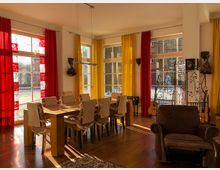 Ein heller Wohnraum mit großen Fenstern, die mit roten und gelben Vorhängen dekoriert sind. Im Zentrum steht ein langer Holztisch mit Stühlen, daneben ein brauner Sessel und ein dekorativer Raumteiler mit verschnörkeltem Metallmuster.