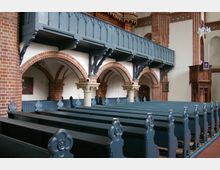 Innenraum einer Kirche mit Sitzbänken aus dunklem Holz in Reihen angeordnet. Im Hintergrund sind rote Backsteinbögen, weiße Wände und eine Empore mit blauen Holzgeländern zu sehen, daneben eine reich verzierte Kanzel aus Holz.