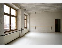 Heller Tanz- oder Übungsraum mit weißen Backsteinwänden, großen Holzrahmenfenstern und einer Ballettstange entlang der Wand. Die Decke besteht aus Holzplanken, und auf dem Boden liegt ein glatter, heller Belag.