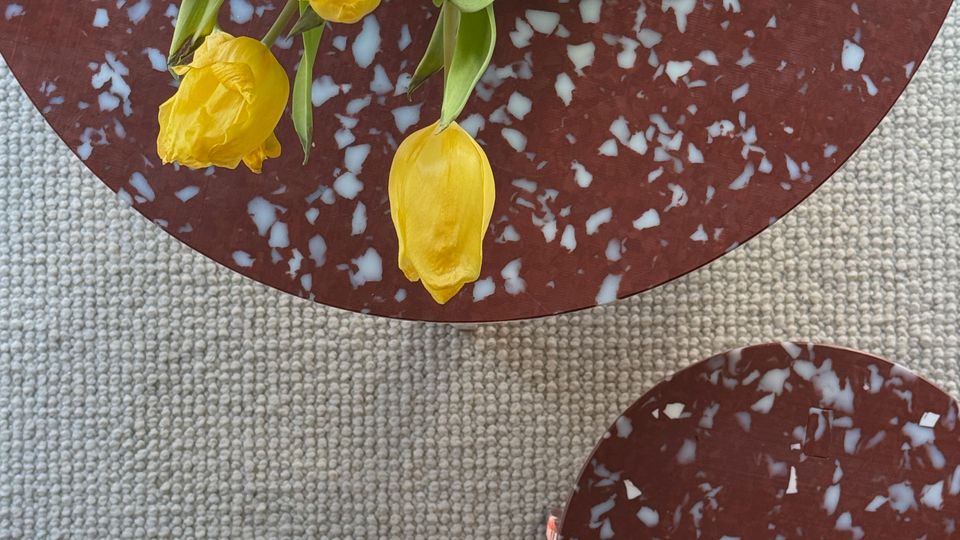 Top-down view of a round, red terrazzo coffee table with white flecks holding a glass vase of yellow tulips, set on a light textured carpet. A smaller matching terrazzo side table sits nearby, with sunlight casting a shadow across the floor.