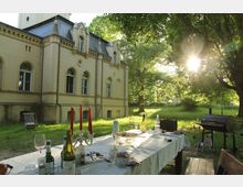 Ein gelber historischer Gebäudeflügel mit verzierten Fenstern steht in einem grasbewachsenen Garten, in dem ein gedeckter Tisch mit Essens- und Getränkeresten sowie ein Grill unter Bäumen im Sonnenlicht zu sehen sind.