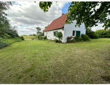 Weiße Landhausfassade mit roten Dachziegeln und grünen Fensterläden, umgeben von einer großen, gepflegten Rasenfläche und dichter Vegetation; im Hintergrund offene Felder unter einem wolkigen Himmel.