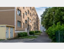 Mehrstöckiges Wohngebäude mit hellbrauner Fassade, flankiert von einem Gehweg auf der rechten Seite, der von Bäumen und Sträuchern gesäumt ist. Im Hintergrund sind weitere ähnliche Wohnhäuser sichtbar.