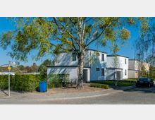 Moderne Wohnhausfassade aus weißem Putz mit flachem Dach, umgeben von einer Hecke und einem Baum im Vordergrund. Das Haus liegt an einer Straßenecke mit Straßenschildern und einem geparkten Auto auf der rechten Seite.