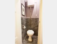 Kleines Badezimmer mit einer weißen Toilette und einem hoch angebrachten Spülkasten, verbunden durch ein Metallrohr. Die Wände und der Boden sind mit dunklen Marmormustern verkleidet, und an der Wand hängt ein gerahmtes Bild.