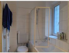 Kleines Badezimmer mit weiß gefliesten Wänden, einer Badewanne mit Duscharmatur und einer gläsernen Duschabtrennung. Neben der Badewanne befindet sich ein Fenster mit Milchglas, und an der Wand hängt ein blaues Handtuch auf einem Handtuchheizkörper.