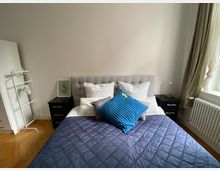 Schlafzimmer mit einem grauen, gepolsterten Bettkopfteil, weißer Bettwäsche und einer blauen gesteppte Tagesdecke. Zwei schwarze Nachttische mit Lampen stehen links und rechts des Bettes, daneben ein weißer Kleiderständer und ein Fenster mit beigefarbenen Vorhängen auf der rechten Seite.