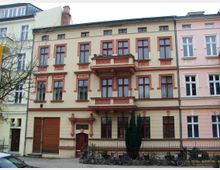 Mehrstöckiges Wohngebäude mit hellbeiger Fassade, rot abgesetzten Fensterrahmen und Balkonen mit dekorativen Verzierungen, flankiert von ähnlichen Bauwerken. Im Vordergrund stehen Fahrräder hinter einem schmiedeeisernen Zaun, und ein kahler Baum ist sichtbar.