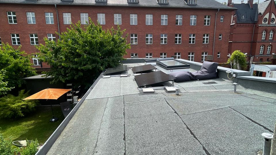 Flachdach mit grauer Bitumenabdichtung, Dachfenstern und niedriger Attika, im Vordergrund mit Sitzkissen. Dahinter ein großer roter Backsteinbau mit vielen weißen Fenstern und Gauben sowie ein begrünter Innenhof unter wolkigem Himmel.