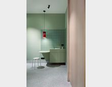 Modernes Badezimmer mit grünen Wänden und Fliesen, einem schwebenden Waschtisch mit Waschbecken in passendem Grünton, roter Pendelleuchte und weißem Hocker auf einem Terrazzo-Boden. Im Vordergrund ist eine Holztür sichtbar.