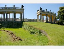 Historische Kolonnaden vor einem grün bewachsenen Hügel, mit Steinmauer und dekorativen Balustraden, Statuen und freistehenden Säulen. Der Himmel ist klar und blau, und die Umgebung wirkt gepflegt.