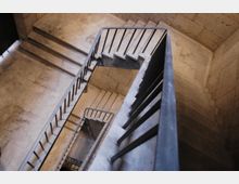 Treppenhaus mit steinernen Treppenstufen und schwarzen, schlichten Metallgeländern, Ansicht von oben in einem Gebäude mit hellen Steinwänden. Die Treppe führt über mehrere Etagen in einem rechteckigen Grundriss nach unten.