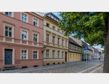 Gepflasterte Straße mit historischen Wohnhäusern in unterschiedlichen Farben, darunter ein rosafarbenes, ein gelbes und ein Fachwerkhaus in Gelb-Braun. Auf der rechten Seite stehen Bäume und Parkschilder, die Umgebung zeigt ruhige, städtische Bebauung.