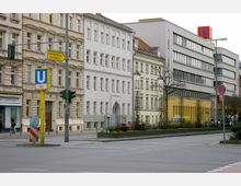 Straßenszene mit mehrstöckigen Wohn- und Bürogebäuden, darunter Altbauten mit weißer und heller Fassade sowie einem modernen Gebäude mit gelbem Erdgeschoss. Im Vordergrund ein Straßenschild für die U-Bahnlinie U9 und Verkehrszeichen, daneben ein Fahrstreifen und karge Straßenbepflanzung.