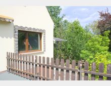Helles Haus mit beigefarbener Fassade, einem Fenster mit Holzrahmen und einer Umrandung aus Ziersteinen. Im Vordergrund steht ein Holzzaun, und im Hintergrund sind Bäume und Sträucher eines Gartens sichtbar.