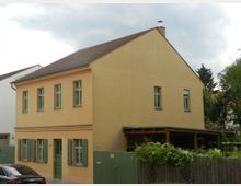 Zweistöckiges Wohnhaus mit gelb-orangefarbener Fassade, grünen Fensterrahmen und Klappläden. Das Gebäude hat ein Schrägdach, einen überdachten Anbau auf der rechten Seite und ist von einem Holzzaun mit Bewuchs umgeben.