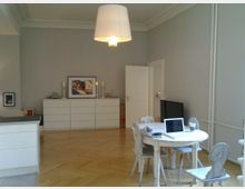 Ein heller Wohnraum mit Parkettboden, grauen Wänden und weißer Deckenleiste. Im Vordergrund steht ein runder Holztisch mit einem aufgeklappten Laptop und vier Stühlen, im Hintergrund befinden sich zwei weiße Kommoden, ein Bild, Kerzenhalter und eine geöffnete Tür.