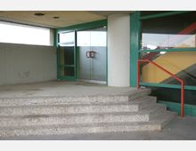 Betonstufen führen zu einem Eingang mit grünen Rahmenelementen und einer verglasten Doppeltür mit roten Griffen. Rechts befindet sich ein rotes Geländer, und die Wand hat Glasfenster mit diagonalen Farbstreifen.