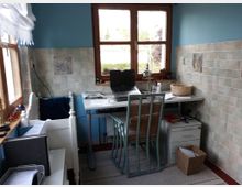 Ein kleiner Arbeitsbereich mit einem weißen Schreibtisch und einem Laptop, umgeben von hellblauen Wänden und pastellfarbenen Fliesen. Zwei Fenster lassen Tageslicht herein, während ein Korbstuhl und diverse Arbeitsmaterialien wie Ordner und Körbe den Raum ergänzen.