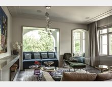 Wohnzimmer mit moderner Einrichtung, darunter eine blaue Sofaecke vor einer großen, bogenförmigen Fensterfront mit Blick auf grüne Bäume. Im Raum befinden sich ein Teppich, Sessel, Couchtische, ein Sideboard und ein Kronleuchter aus Glas.