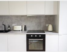 Moderne Küche mit weißen Schränken, grauer Arbeitsplatte und Wand in Betonoptik. Sichtbar sind ein schwarzes Spülbecken mit Wasserhahn, ein cremefarbener Toaster, ein Wasserkocher und ein eingebauter Backofen unter einem Induktionskochfeld.