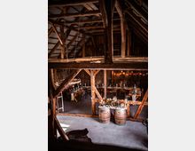 Innenraum einer rustikalen Scheune mit Holzbalken und einer hohen Decke. Im Vordergrund stehen dekorierte Weinfässer, während im Hintergrund gedeckte Holztische mit Stühlen für eine Veranstaltung arrangiert sind.