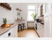 Helle, moderne Küche mit weißen Schränken, einer Arbeitsplatte aus Holz und einem Fenster, das Tageslicht hereinlässt. Auf der linken Seite steht ein Tresen mit zwei Hockern, während auf der rechten Seite ein Ofen, ein Herd und Küchengeräte zu sehen sind; eine Schale mit Obst sowie eine Zimmerpflanze verleihen dem Raum dekorative Akzente.