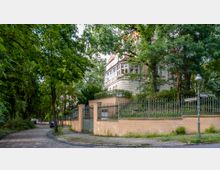 Eine freistehende, historische Villa mit heller Fassade und einem Erker, umgeben von einem Metallzaun auf einer kleinen Mauer. Die Straße ist mit Kopfsteinpflaster bedeckt, und der Bereich ist von zahlreichen Bäumen und Grünflächen geprägt.