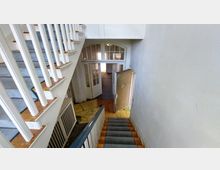 Treppenhaus einer älteren Gebäudeinnenansicht mit weiß gestrichenen Holzgeländern, einer Holztreppe und einer halboffenen Holztür. Im Hintergrund führen verglaste Doppeltüren zu einem beleuchteten Raum.