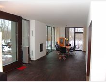 Innenraum eines modernen Wohnhauses mit dunklem Holzboden, großen Glasschiebetüren und bodentiefen Fenstern, die den Blick auf einen verschneiten Außenbereich freigeben. Ein Esstisch mit schwarzen Stühlen steht im Hintergrund, daneben ein orangenes Schrankmöbel.