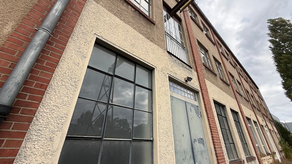 Schräg aufgenommene Fassade eines mehrstöckigen Industrie- oder Lagergebäudes mit rauem Putz, roten Ziegelpfeilern und großen, schwarz gerahmten Sprossenfenstern. Neben einem verwitterten Metalltor sind Regenfallrohre und eine kleine Außenlampe zu sehen, im Hintergrund Himmel und Bäume.